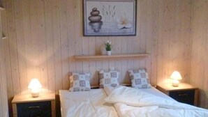 Une chambre avec un mur lambrissé en bois, un lit avec une literie blanche, deux tables de chevet avec des lampes et une petite étagère avec une plante.