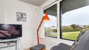 Ett vardagsrum med en tv, en grå fåtölj, en liten pall, en röd golvlampa och ett fönster med utsikt över omgivningen.