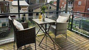 A balcony with wicker chairs and a table, overlooking a street with buildings and a parked car. - Ferienwohnung Sturmeck, Fehmarn-orth (Fehmarn)