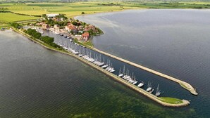 An aerial view of a marina with boats docked along a narrow pier, surrounded by green fields and a small cluster of buildings. - Ferienwohnung Sturmeck, Fehmarn-orth (Fehmarn)