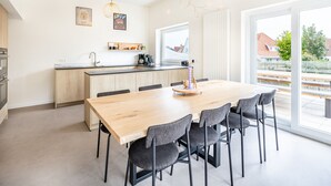 A modern kitchen with a central island, a dining area with a wooden table and chairs, and large windows offering a view of the outside. - Villa in Koksijde Near the Sea (Koksijde)
