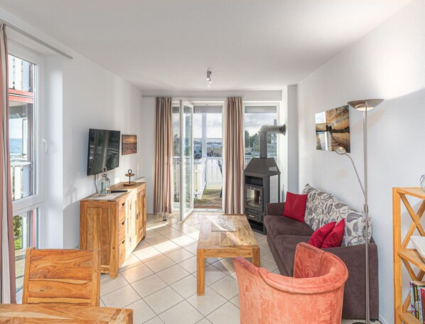 A modern living room with a fireplace, wooden furniture, and a large window offering a view of the outdoors. - Appartement im Ferienpark in Rechlin (Rechlin)