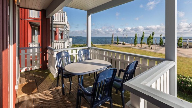 A covered outdoor seating area with a table and chairs, overlooking a body of water and a grassy area with trees and flags. - Appartement im Ferienpark in Rechlin (Rechlin)