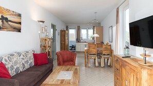A living room with a sofa, coffee table, and a TV. There is a dining area with a table and chairs. The room has a kitchen in the background. - Appartement im Ferienpark in Rechlin (Rechlin)