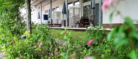 A covered outdoor seating area with chairs and umbrellas, surrounded by lush greenery and flowering plants.