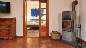 A room with a wooden dresser, a television, a bed with a blue abstract painting, a stone wood stove, and a wicker basket. - Ferienhaus Oliver, Wendorf (Kuhlen-Wendorf)