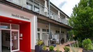 Entrada de un hotel con toldo rojo, mesas al aire libre y plantas en macetas.