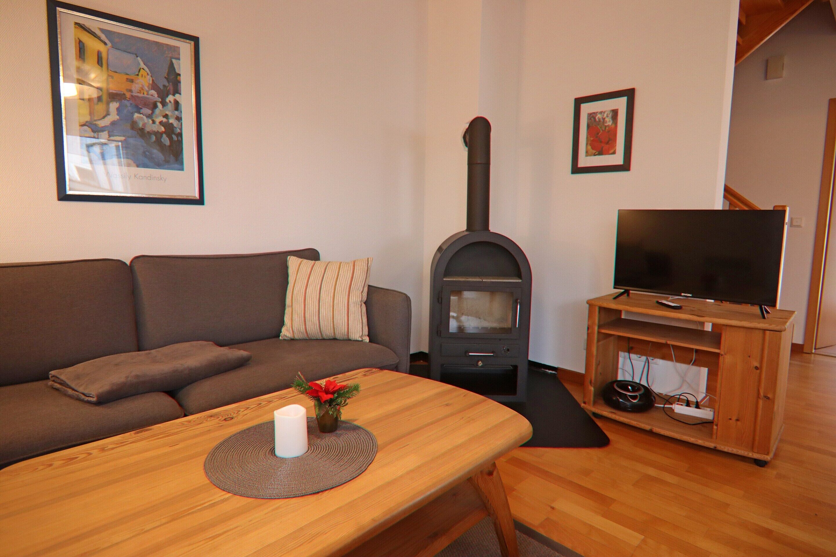 Ein Wohnzimmer mit einer Couch, einem Holztisch mit Kerze und Pflanze, einem Kaminofen und einem Fernseher auf einem Holzschrank.