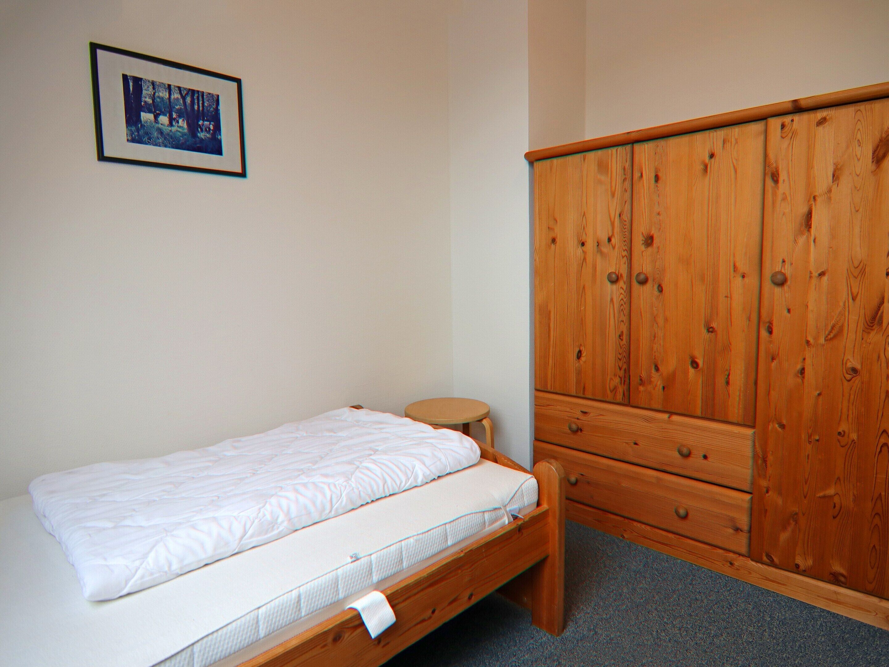A single bed with a wooden frame and a mattress, a small round stool, and a wooden wardrobe with multiple drawers.
