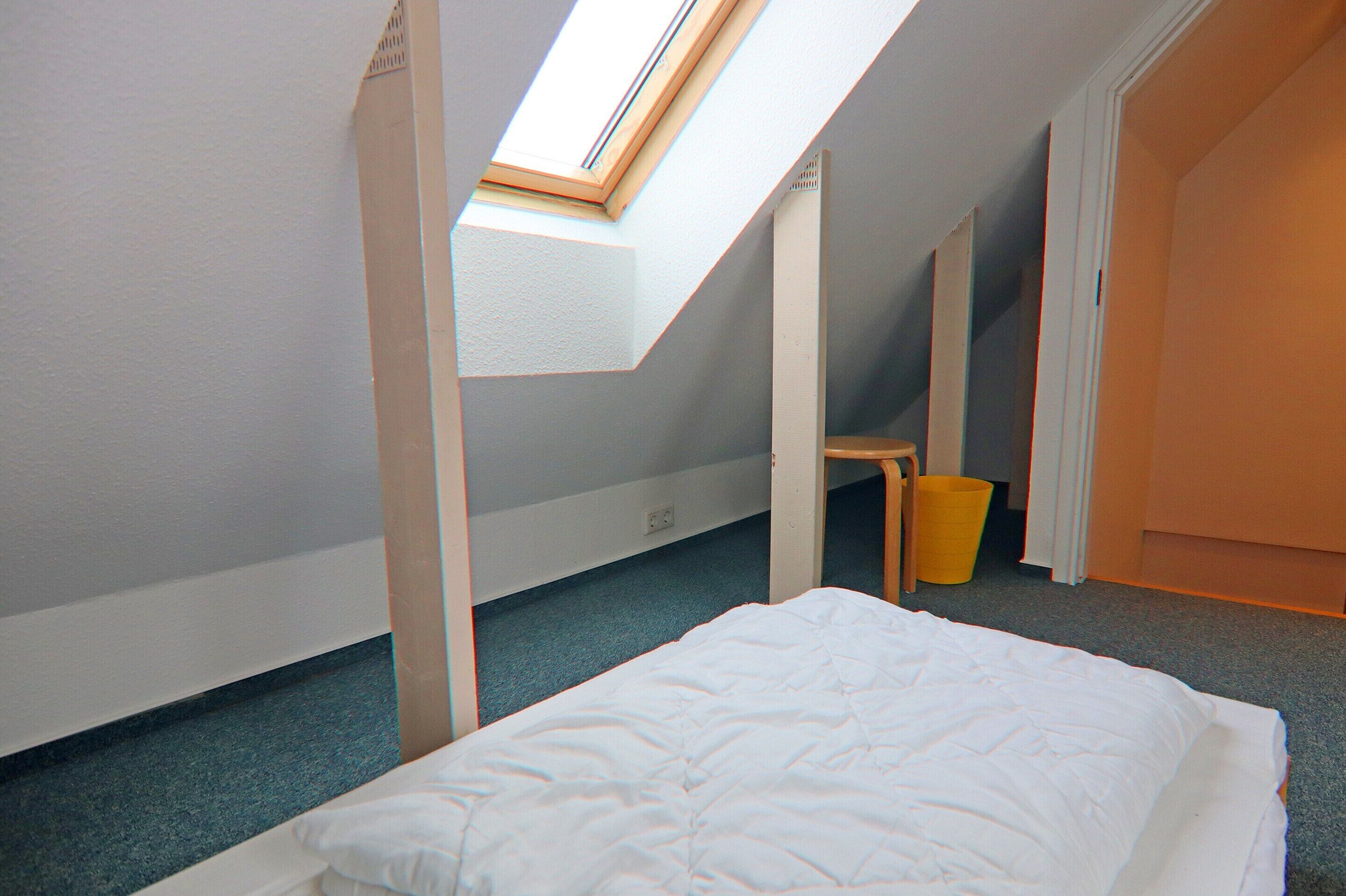 A room with a skylight, a bed, a chair, and a trash bin.