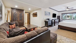 A modern living room with a sofa, a coffee table, and a TV. There is a bed with a window in the background.