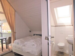 Ein Schlafzimmer mit Bett, einem Sessel, einem Badezimmer mit Toilette und Waschbecken sowie einem Balkon mit Vorhängen.