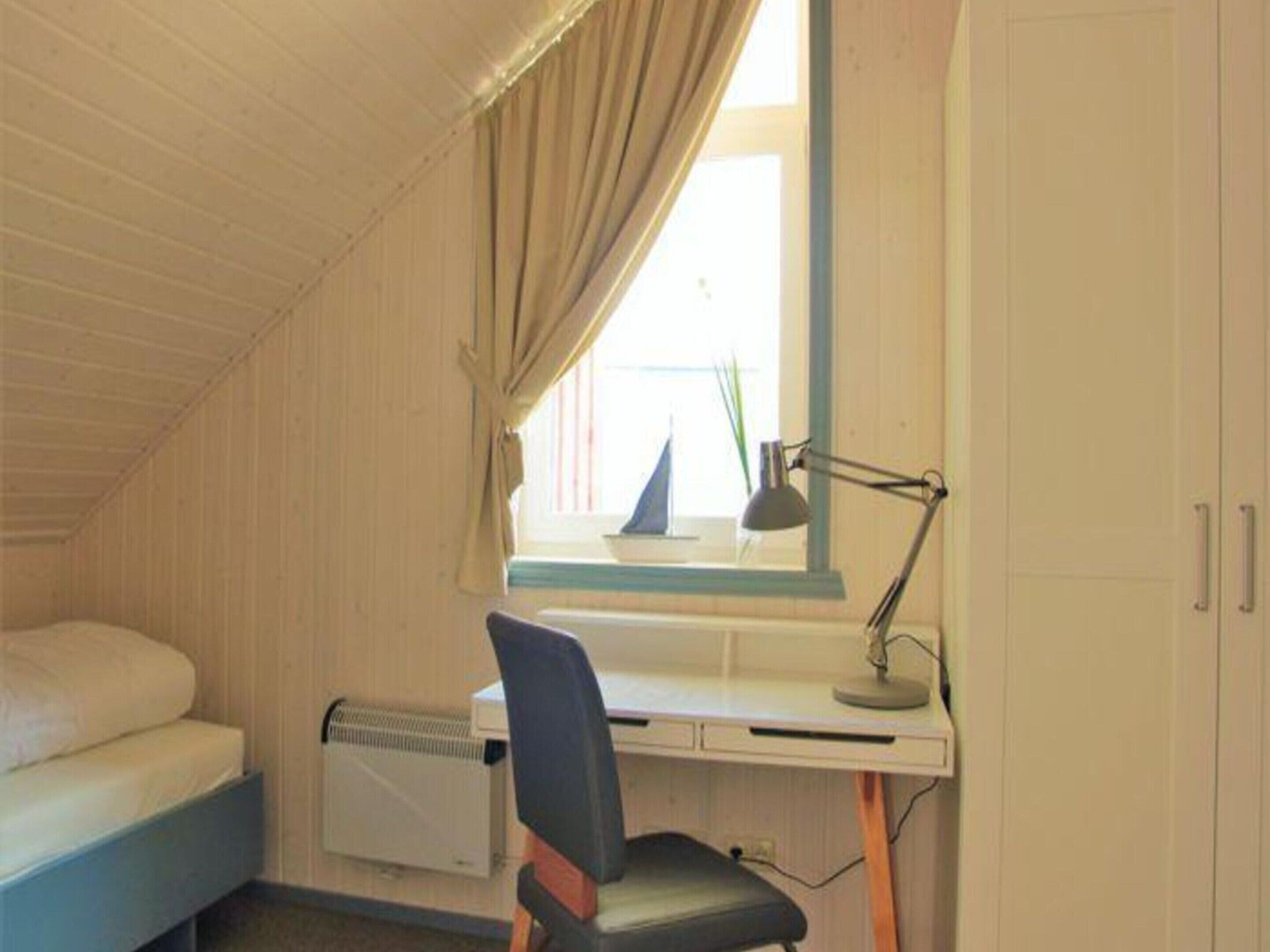 A dormitory room with a bed, desk, chair, radiator, and a window with curtains.