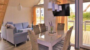 A modern living room with a sofa, a dining table with chairs, a flat-screen TV mounted on the wall, and large windows offering a view of the outdoors.
