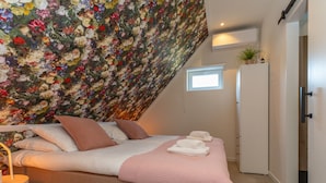 A bedroom with a sloped ceiling, a bed with pillows and a pink blanket, a bedside table with a lamp, and a small window. - Apartment in Kortgene Near Grevelingenmeer (Kortgene)