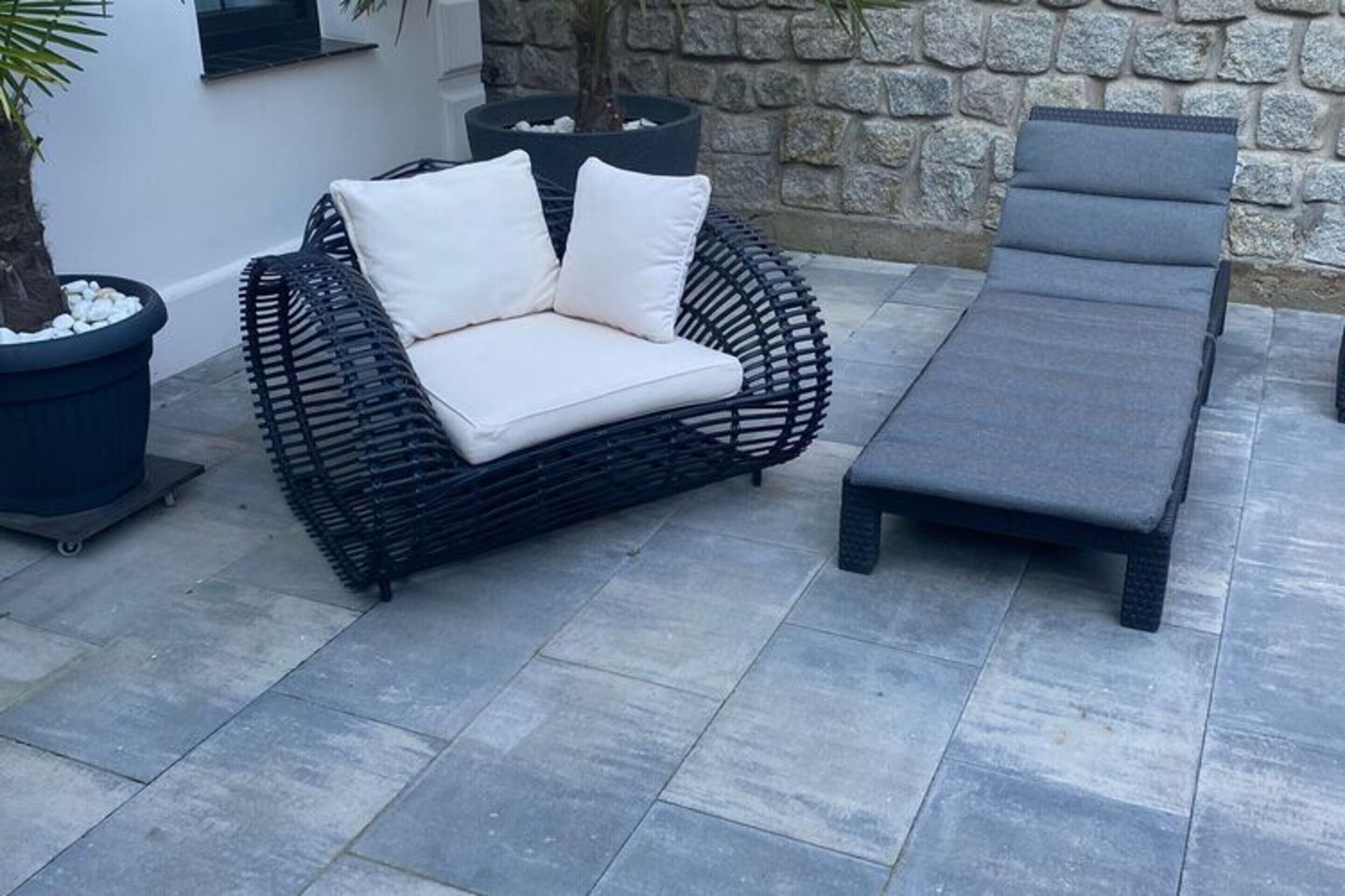 A wicker sofa with white cushions and a grey lounge chair on a tiled patio.