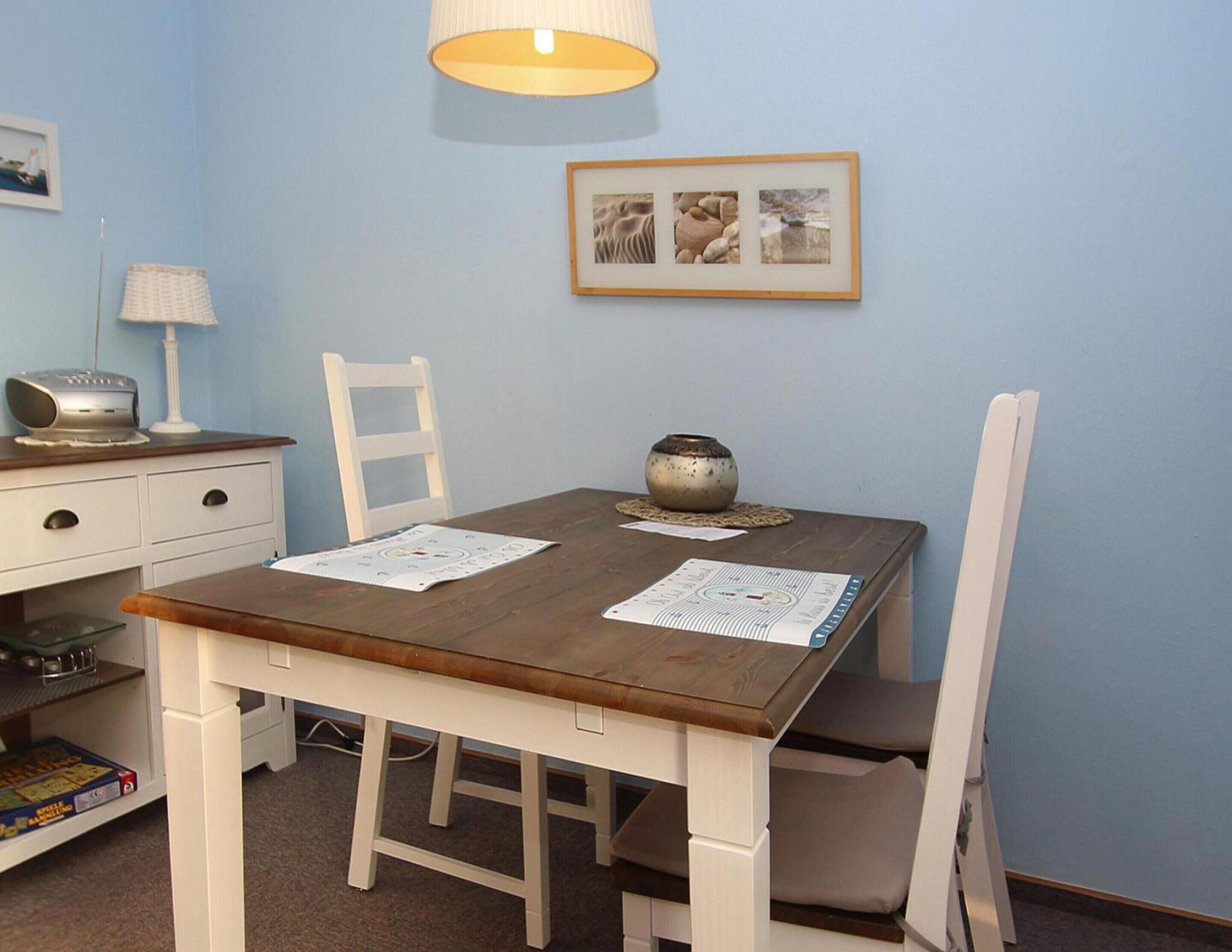 A dining area with a wooden table, white chairs, a lamp, a framed picture, and a small table with a bowl.