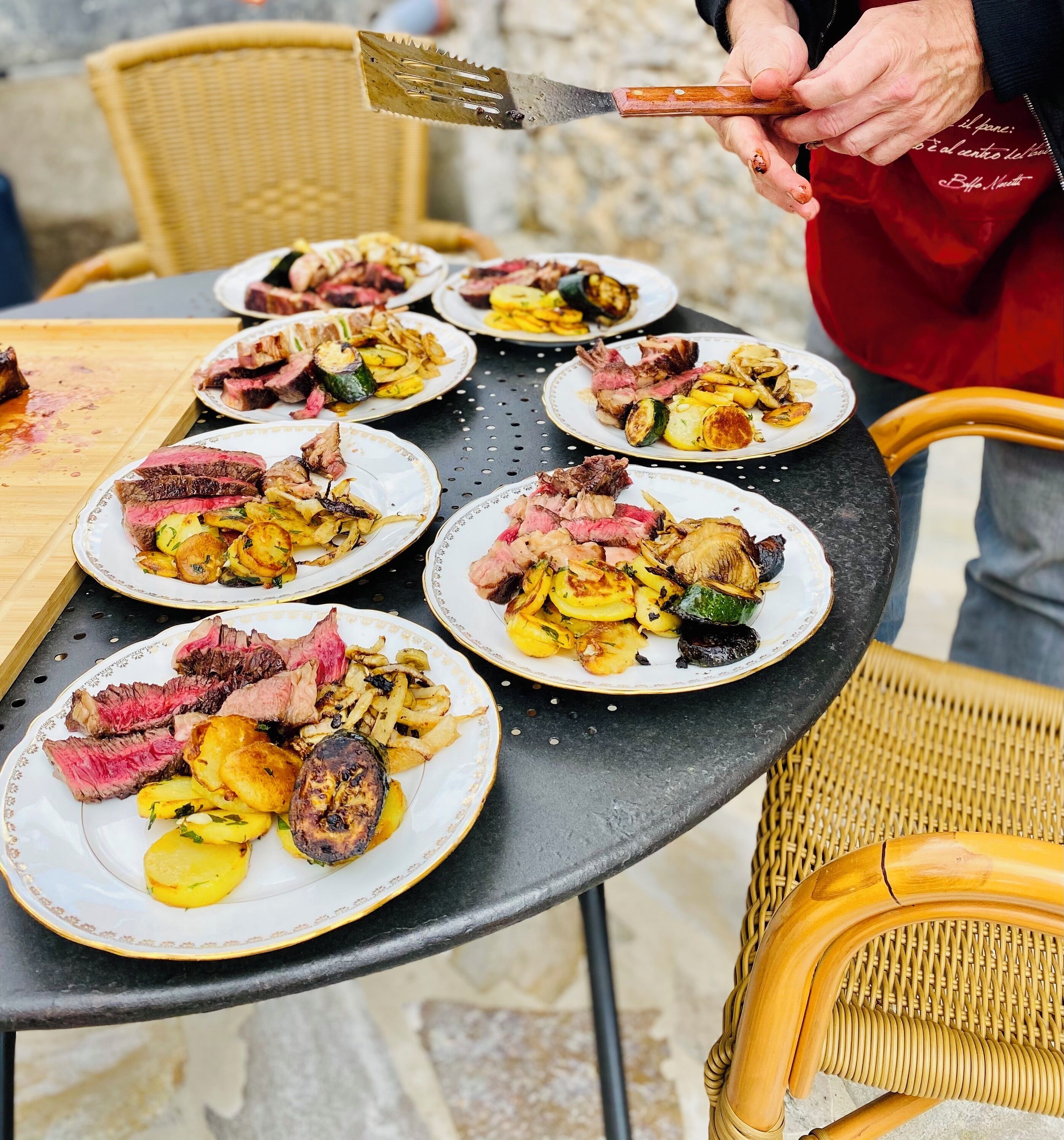 A dining table with plates of food, a person holding a spatula, and wicker chairs.