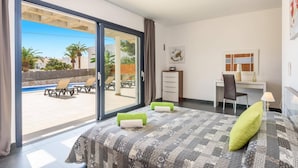 A hotel room with a large sliding glass door leading to an outdoor pool area, a bed with patterned bedding, a desk with a chair, and a small table with a lamp. - Villa Primasol by Villa Plus (Punta Prima)