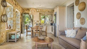 A rustic kitchen with stone walls, a wooden dining table, and a cozy living area with a sofa and cushions. - Villa Kalou Cottage by Villa Plus (Ionian Islands)