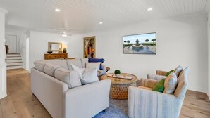 Ein geräumiges Wohnzimmer mit einem weißen Sofa, einem gestreiften Sessel und einem Couchtisch. Ein Bildbrunnen ist an der Wand zu sehen, und im Hintergrund ist eine Treppe erkennbar.