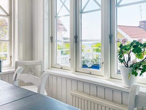 A room with a table and chairs, large windows with white frames, and a view of greenery and buildings outside. - 4 Star Holiday Home in Kyrkesund (Kyrkesund)