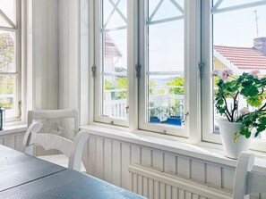 Una habitación con una mesa y sillas, ventanas amplias con marcos blancos y vista a una cerca blanca y follaje exterior.