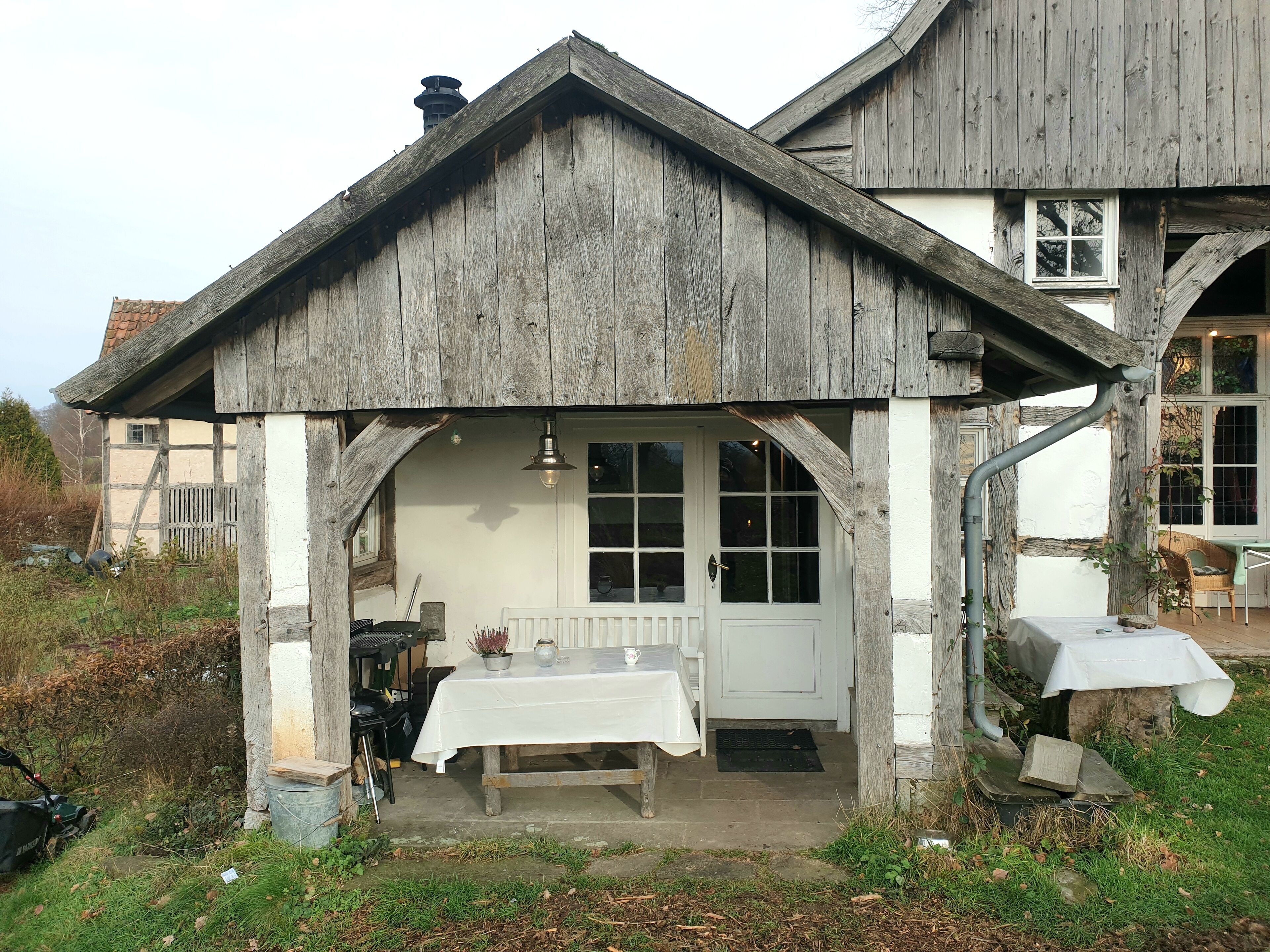Una construcción rústica de madera con puerta y ventanas blancas, una mesa con mantel blanco y una silla afuera.