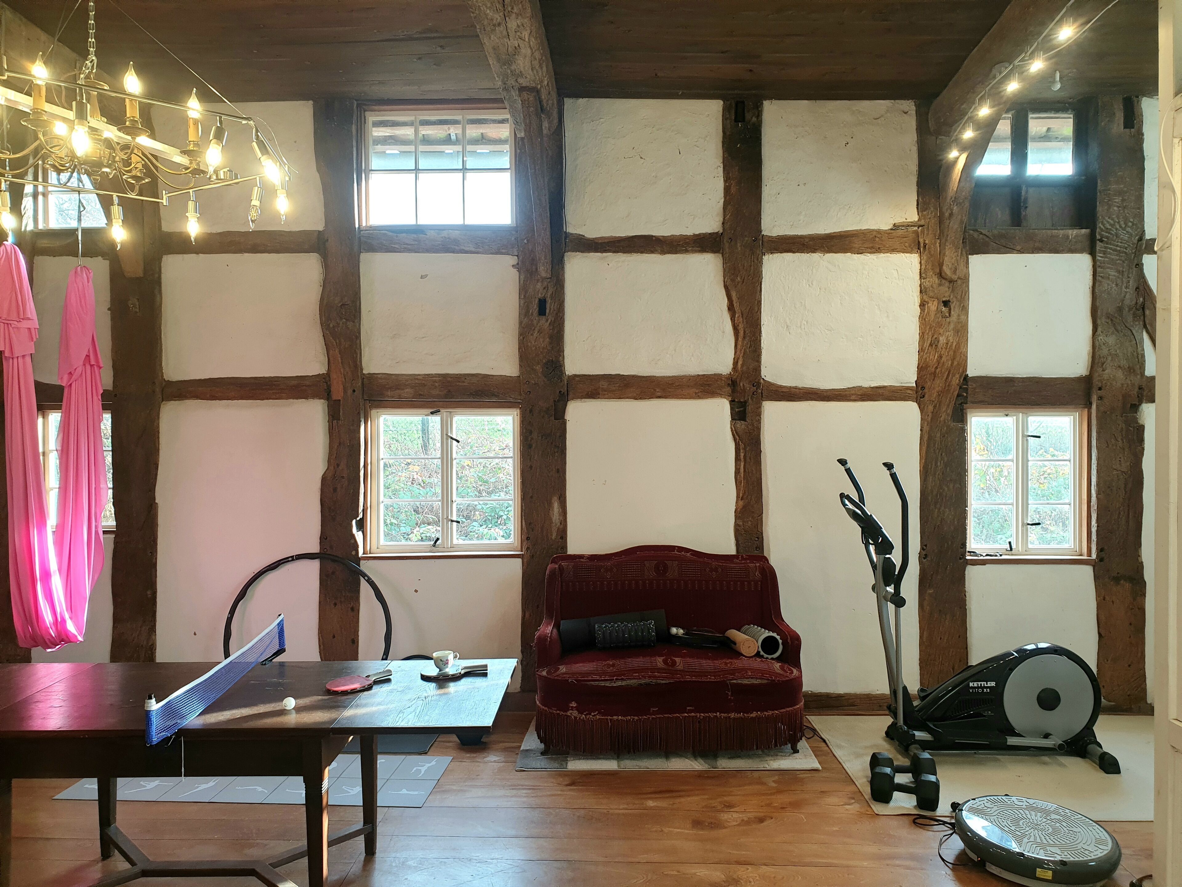 Ein Zimmer mit freiliegenden Holzbalken, einem roten Sofa, einem Tisch mit Trainingsgeräten und großen Fenstern mit Blick auf Grünflächen.