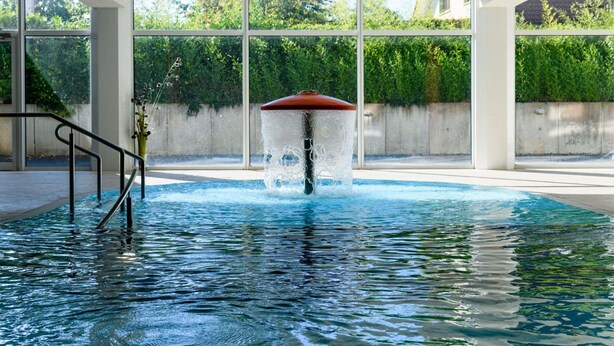 Ein Schwimmbecken mit einer Fontäne, umgeben von großen Fenstern und einer Betonmauer.