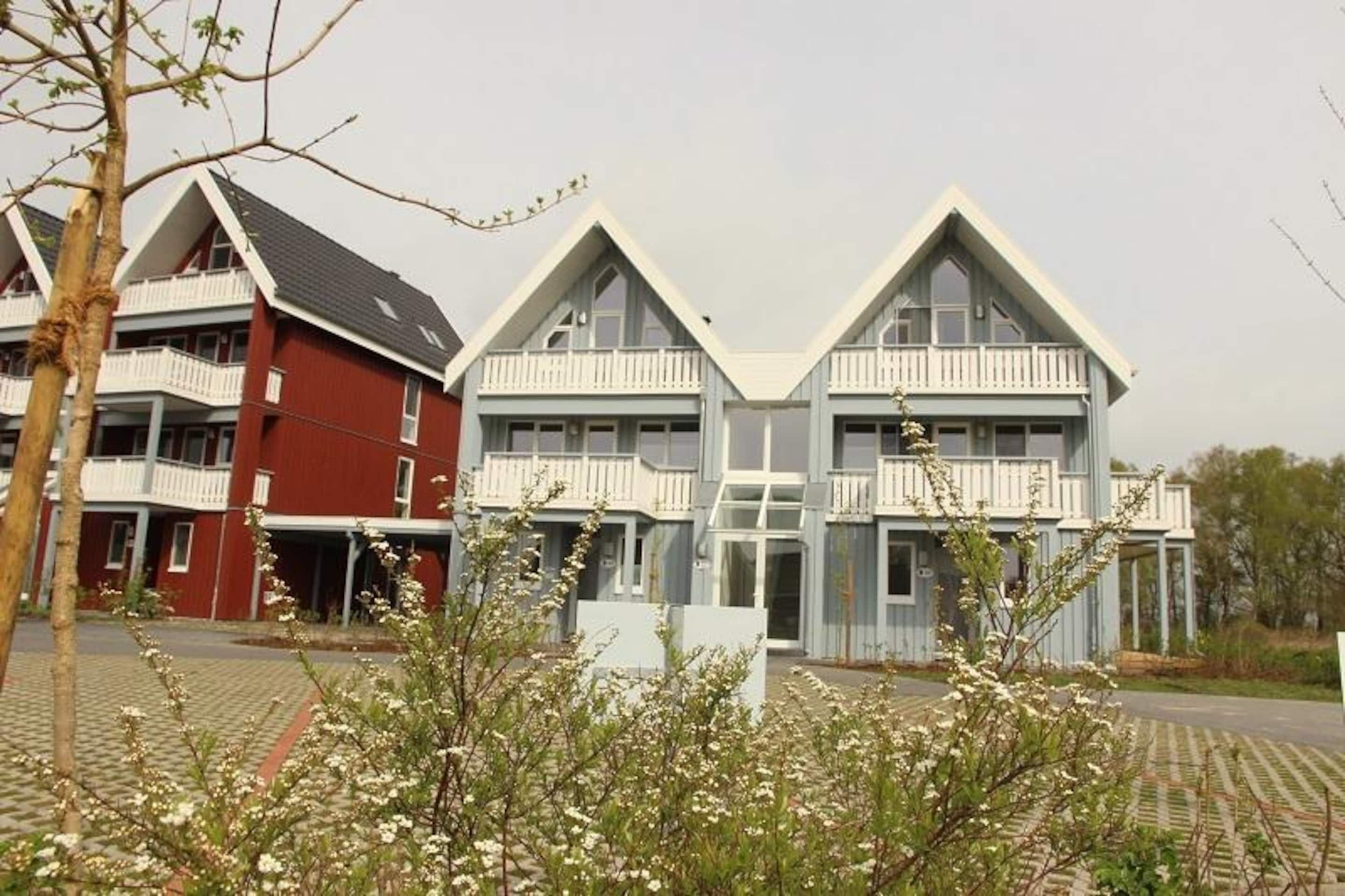 A row of two-story houses with balconies, a paved driveway, and flowering bushes in the foreground.