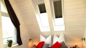 A bedroom with a sloped ceiling, two windows with blinds, a bed with grey and red pillows, a bedside table with a lamp, and a small plant on the window sill. - Liebevoll Eingerichtetes Appartement (Rechlin)
