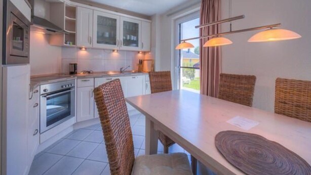 Une cuisine moderne avec des meubles blancs, un four encastré et un coin repas agrémenté de chaises en osier et d’une longue table.