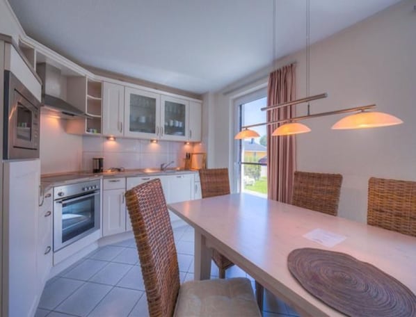 A modern kitchen with white cabinets, a built-in oven, and a dining area with wicker chairs and a long table.