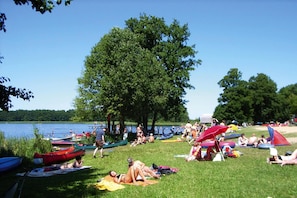A lakeside picnic area with people relaxing on the grass, boats, and umbrellas. - Apartment in Granzow Near the Lake (Mirow)