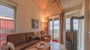 A cozy living room with a sofa, armchair, and a table with placemats. A wood-burning stove is visible, and there are large windows with curtains. - Appartement im Ferienpark in Rechlin (Rechlin)
