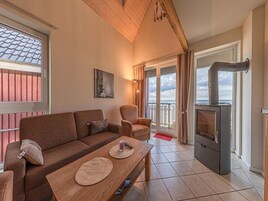 Una sala de estar acogedora con un sofá, un sillón y una mesa con individuales. Se ve un estufa de leña, y hay ventanas grandes con cortinas.