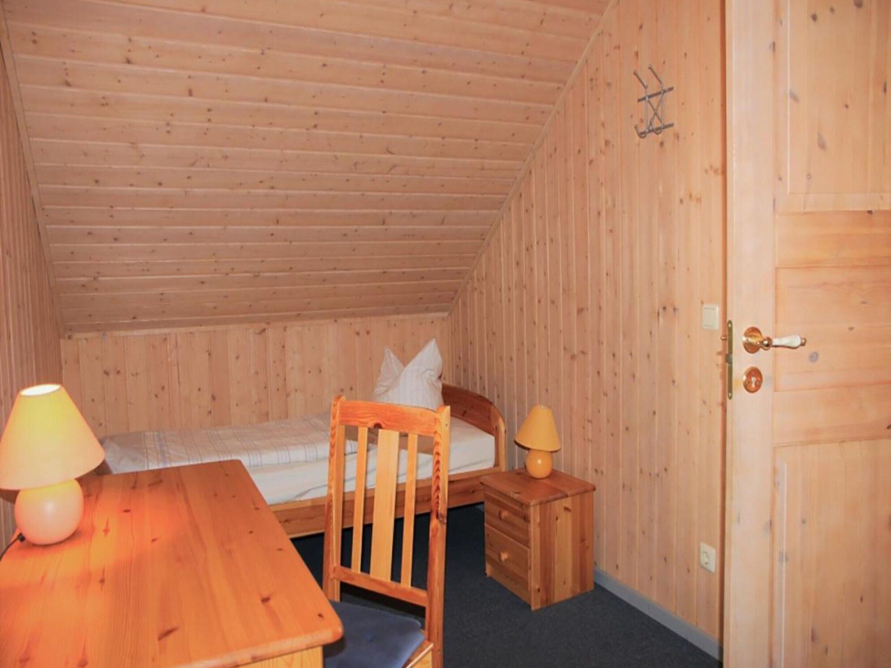 Une petite pièce aux murs lambrissés, comprenant un lit simple, un bureau en bois et une chaise.