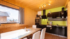 Interior de madeira com uma mesa de jantar e cadeiras, cozinha com armários pretos e uma parede verde, e uma janela com cortinas.