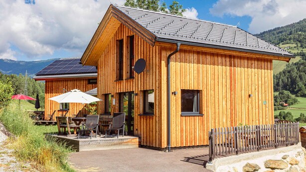 Uma cabana de madeira com telhado inclinado, painéis solares e um alpendre com área de estar ao ar livre.