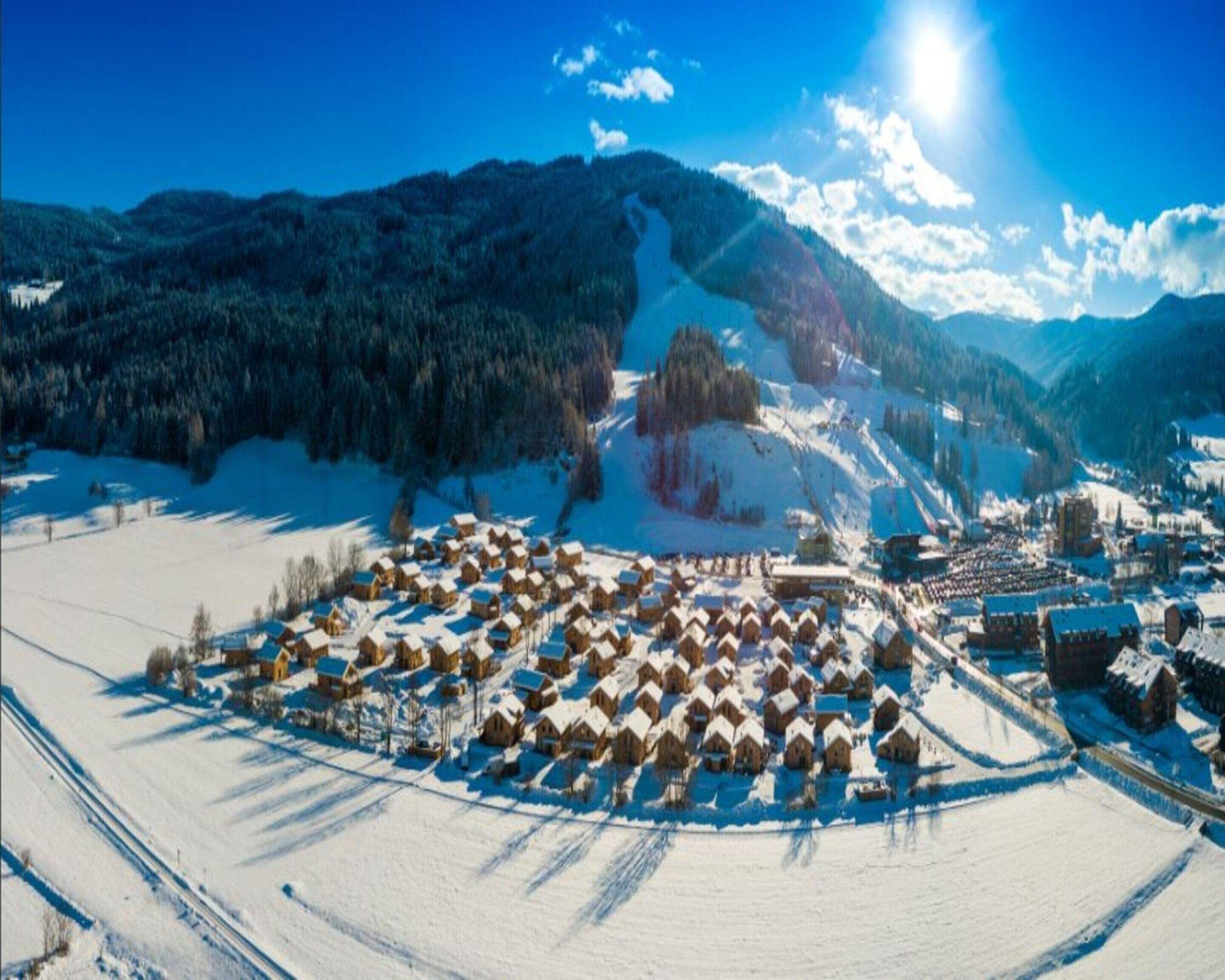Ein schneebedecktes Dorf mit Holzhäusern, die in einer Ringform angeordnet sind, umgeben von Bergen und einem klaren Himmel.