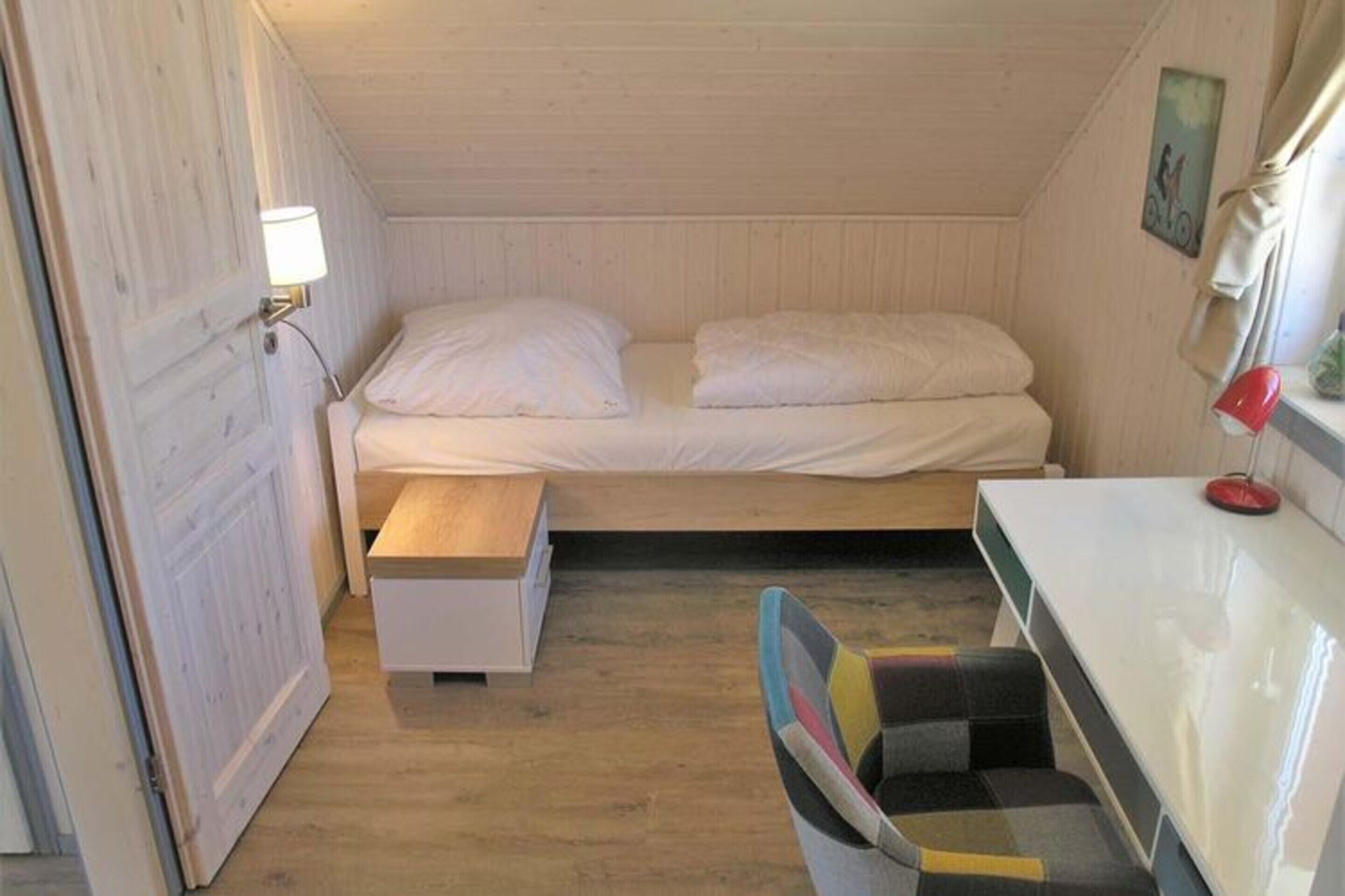 A small, cozy room with a bed, a bedside table, a chair, and a small desk with a lamp.
