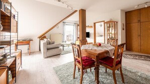 Een eetgedeelte met een houten tafel, stoelen en een rode gewatteerde bank. Links is een trap met houten leuning te zien, en er staat een bankstel op de achtergrond.