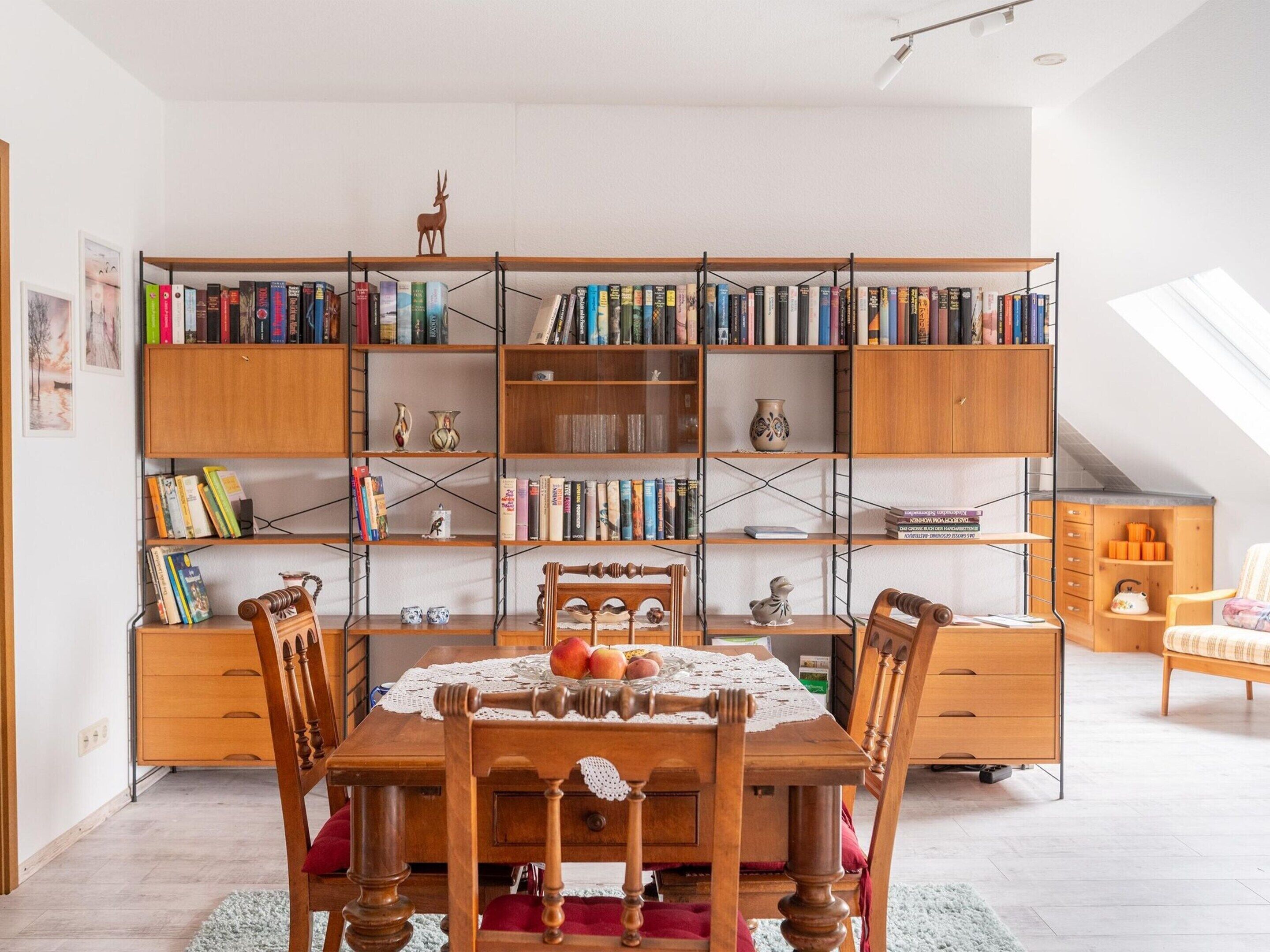 A cozy room with a wooden bookshelf filled with books, a dining table with chairs, and a small wooden cabinet with a kettle.