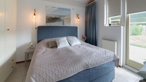 A bedroom with a large bed, a headboard, a nightstand, a wall-mounted lamp, a window with curtains, and a painting on the wall. - Spacious Holiday Home With Sauna Near Beach (Wemeldinge)