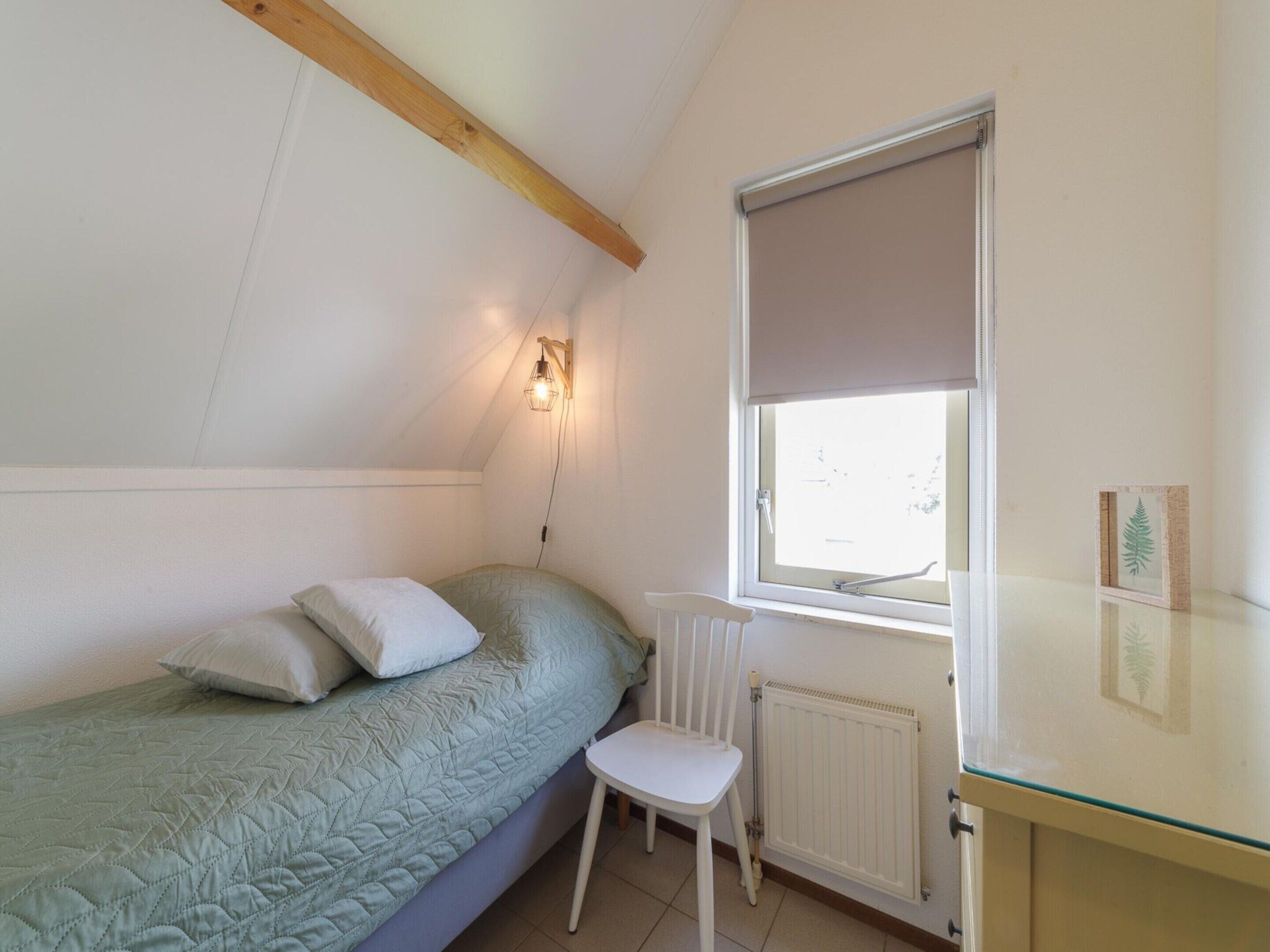 Une petite chambre avec un lit simple, un plafond en pente, un radiateur, une fenêtre avec un store et une petite table avec une chaise.