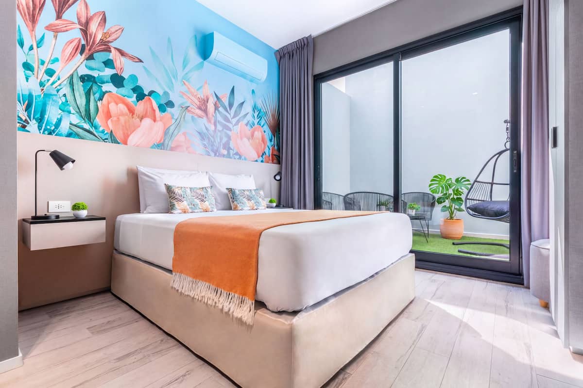 A modern bedroom with a large bed, a nightstand, a wall-mounted air conditioner, and a sliding glass door leading to a balcony with plants.