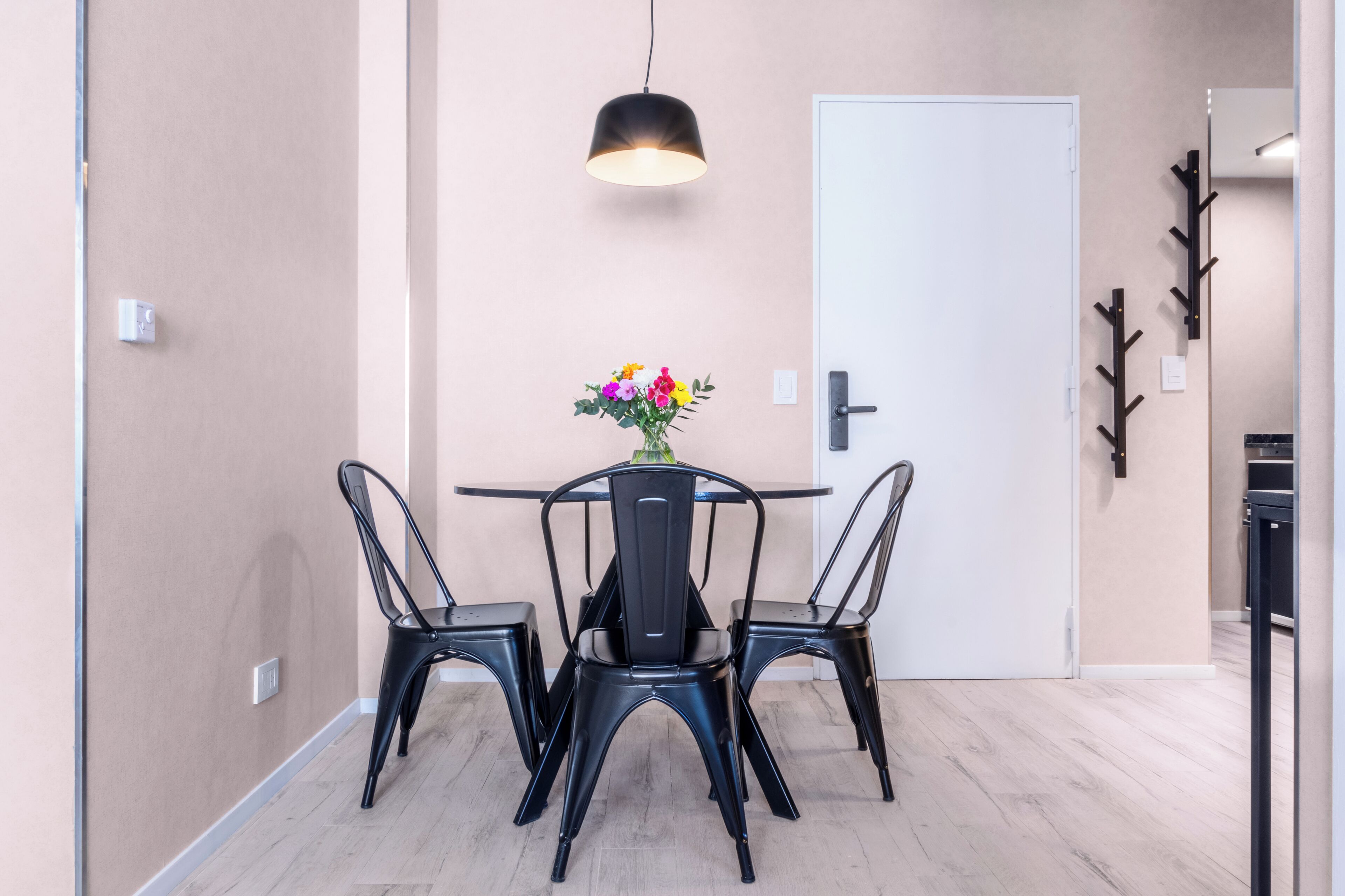 A dining area with a table and four chairs, a vase with flowers, a hanging light, and a door with a coat rack.