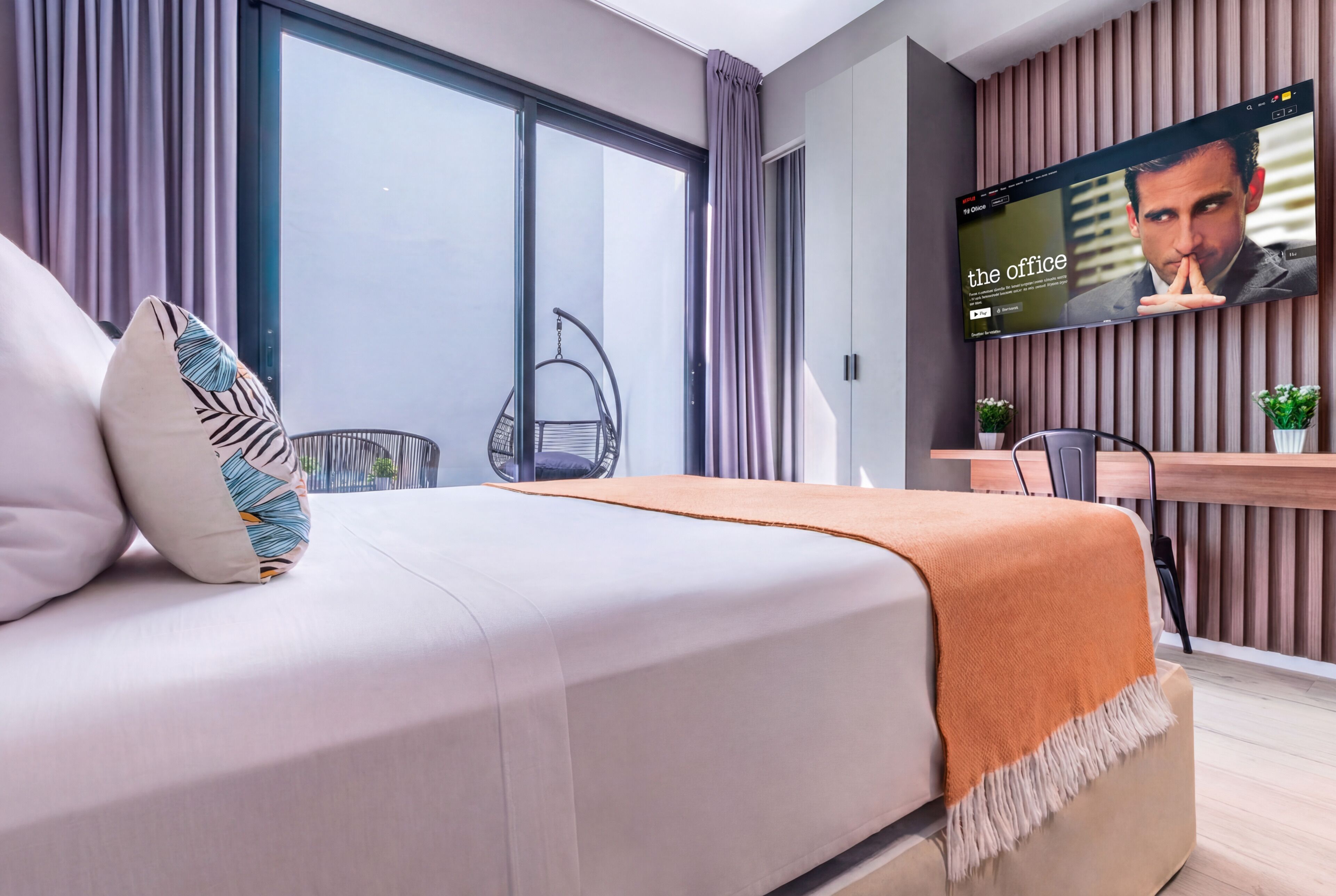 A modern hotel room with a bed, a TV screen displaying 'The Office', a balcony with a chair, and a wooden desk.