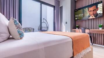 A modern hotel room with a bed, a TV screen displaying 'The Office', a balcony with a chair, and a wooden desk.